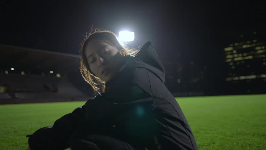 A woman kneeling on an athletics field at night rests for a moment before being approached by a man and helped to her feet