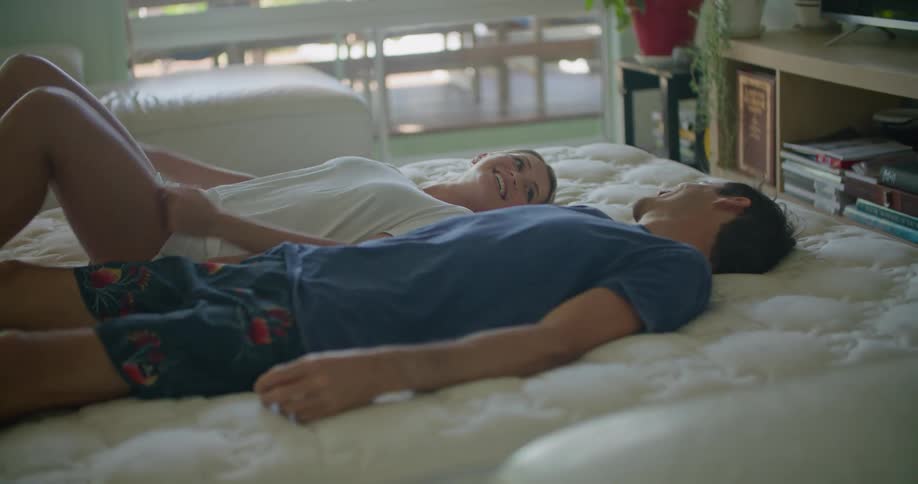 A man and woman lay down on a bed while smiling and holding hands