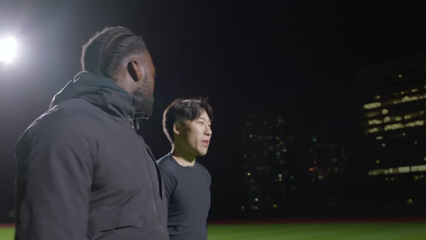 A pair of men walking across a sports field at night talk with one another before breaking out into a jog