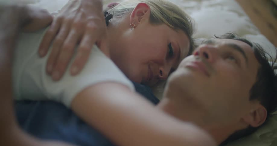 A man and woman embracing while lying on a bed look at one another and smile