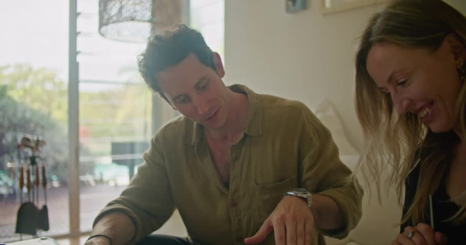 Man with yellow shirt and black pants and blonde woman wearing black blouse sit on beige couch looking down at documents talking to each other and smiling in living room at home
