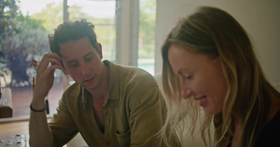 A man and woman discuss the contents of a notepad while sitting together on a sofa indoors during the day