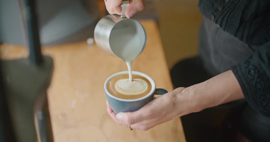 Someone is holding a cup of coffee in one hand and pouring cream into it with the other hand