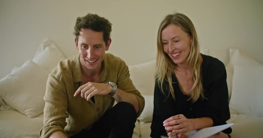 A man and a woman sit on a couch laughing at something they are watching on the laptop