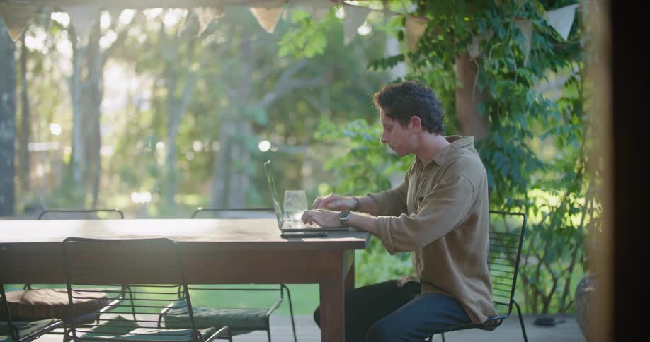 A man typing on a laptop is distracted for a moment by a dog playing with a ball before returning to his typing