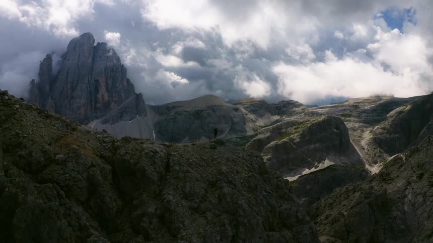 A bird flies over a person standing on top of a mountain ridge and there are more mountains in the distance