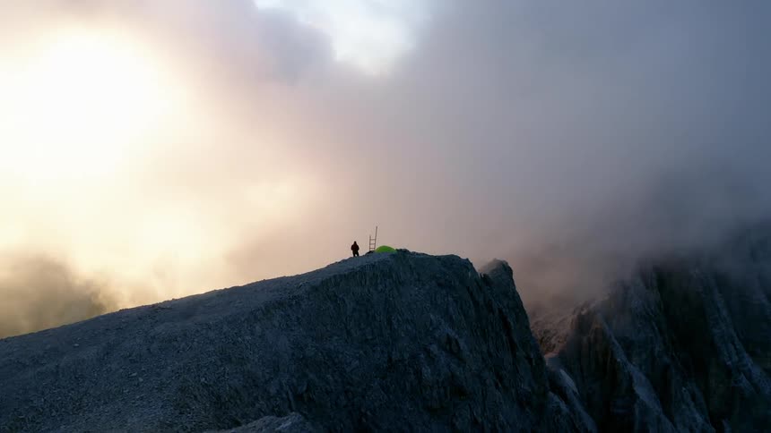 Person stands near tent placed on plateau near ice patch overlooking sheer cliff among low lying clouds