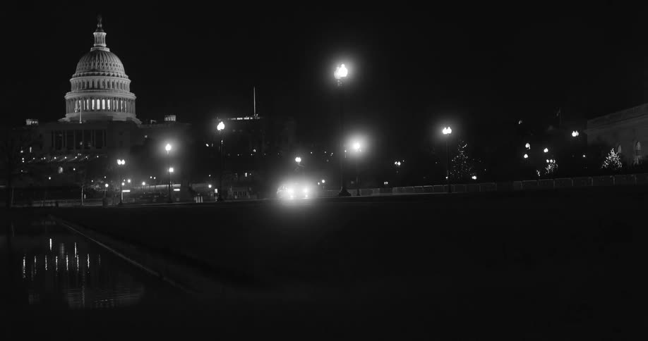 Car drives along road away from capitol building passing pond at night