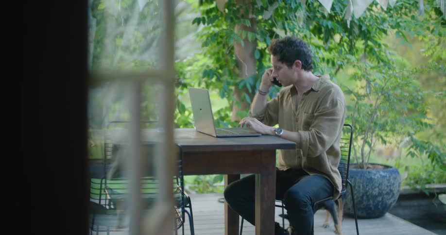 A man is sitting at a table outside talking on the phone and typing on his laptop computer as a dog plays near him