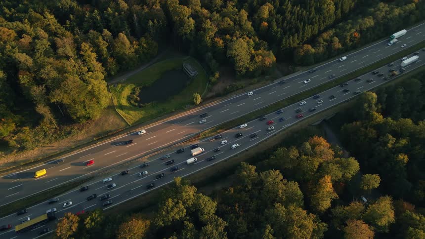 Traffic flows slowly along one side of divided highway passing over bridge and through forest with trees starting to change to autumn colors
