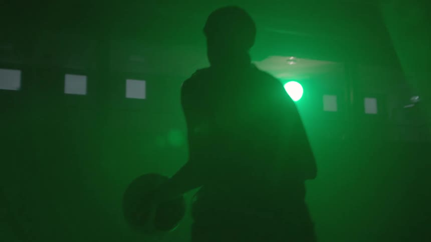 A spotlight is shining on the dark court as a woman dribbles a basketball
