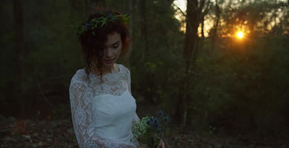 Woman poses in wedding dress near woods as sun shines through trees behind her
