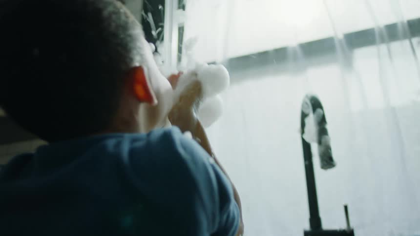 A boy and girl on handfuls of soap suds while standing at the kitchen sink
