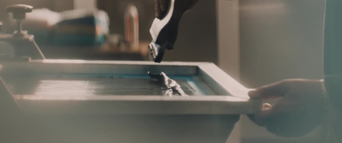 Person spreads paint across silkscreen then silkscreens pattern onto t shirt in workshop