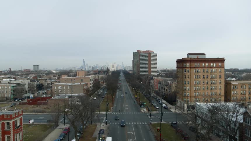 Traffic flows down leafless tree lined street through residential area toward city skyline
