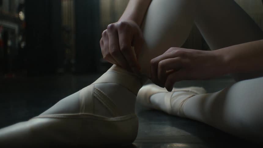 A ballet dancer is finishing tying a knot in the strapping of her ballet shoes while sitting on the floor in a room