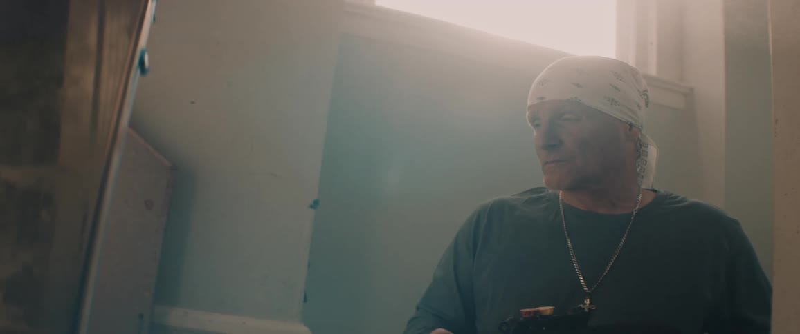 A man wearing a white bandanna is sitting down using a tool and smoke is rising up to the light shining in from the window