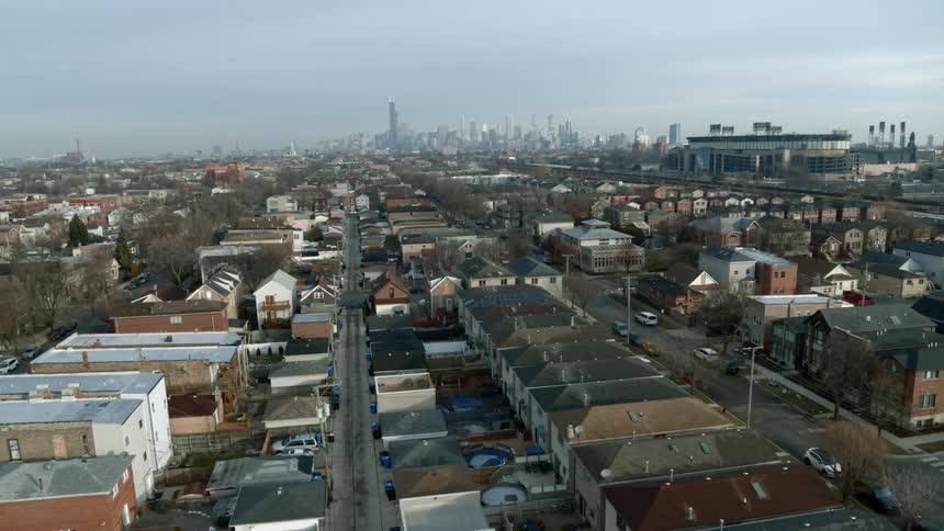 An aerial view of houses and cars lining streets in a neighborhood with the downtown skyline in the far distance