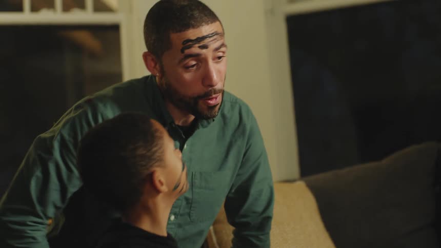 A boy applies dark finger paint to his dads face as his mom cheers them on