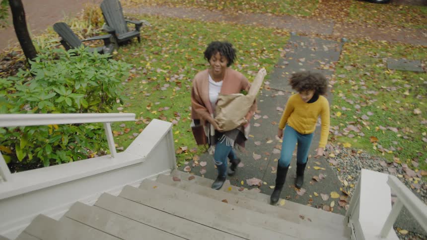 Fruit spills out from the bottom of a grocery bag as a woman ascends a staircase with her daughter