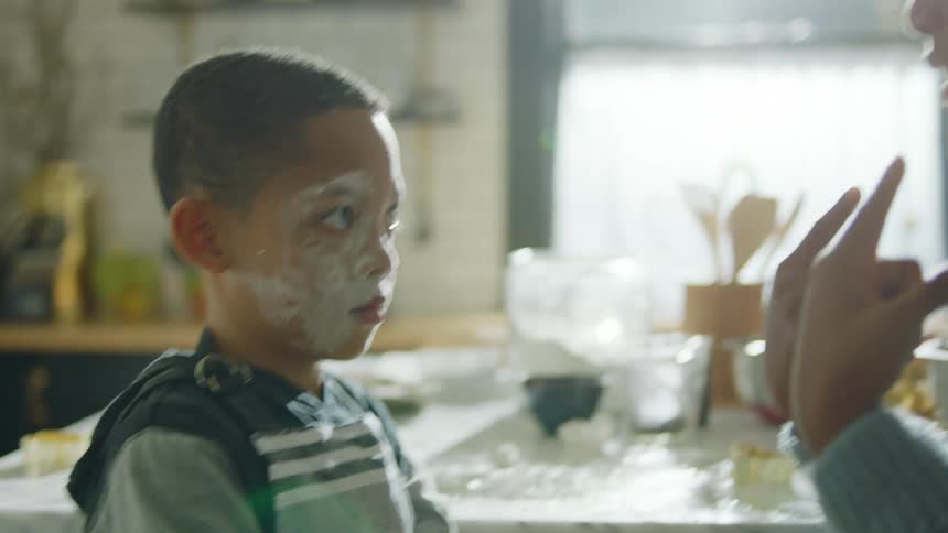 Mother scolds son while wiping at his flour covered face while they stand near messy kitchen island