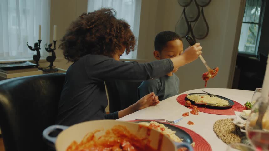 Sister serves pasta sauce onto brothers spaghetti spilling some on tablecloth