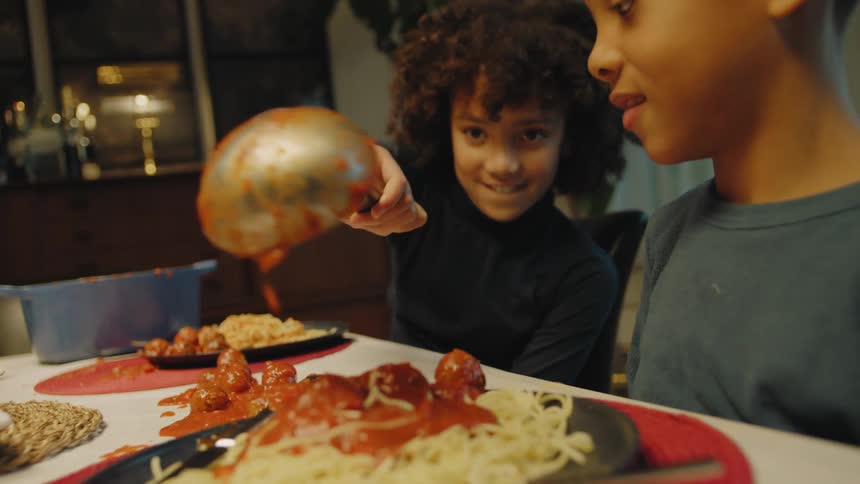 A young girl sits at the dining table at home and takes a scoop of meatballs and sauce out of a pot and dumps in on the table instead of her brothers plate