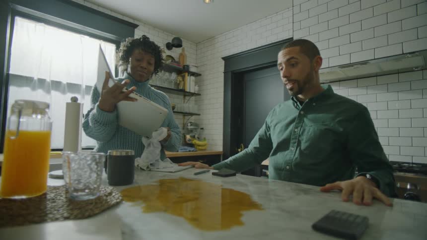 A woman is standing in a kitchen holding a laptop computer and wiping it with a towel as a man is sitting down with a spilled drink on the counter in front of him