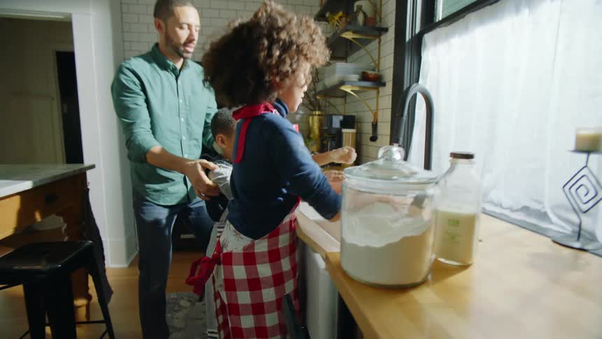 A girl washing her hands in the kitchen sink pouts when her parents carry her brother to the sink to wash his hands as well