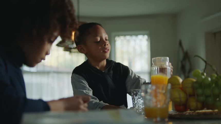 A boy prepares to pour himself a glass of orange juice while sitting at a table next to his studying sister