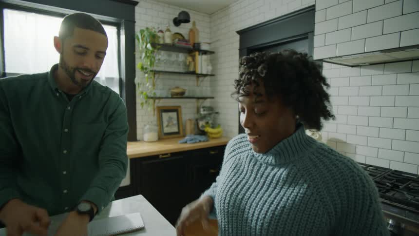 A woman is sitting at a kitchen table talking to a man and he hugs and kisses her