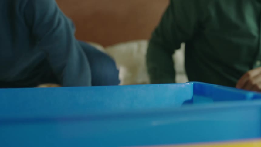 A boy and his family sit in bed and throw small toys into a bin while cleaning