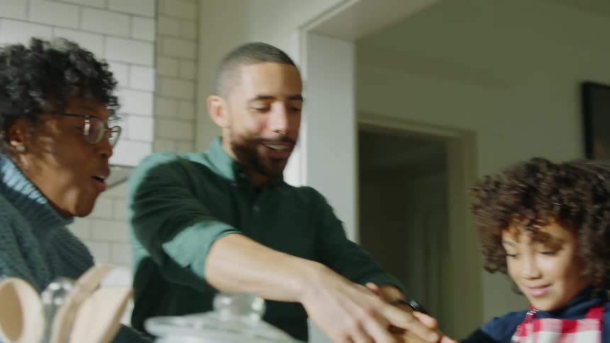 Son spills cup of milk handed to him by mother while family cooks together in kitchen