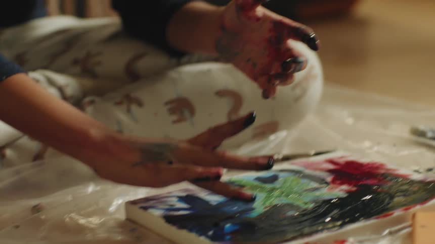 Person sits cross legged on plastic sheet covered wooden floor finger painting