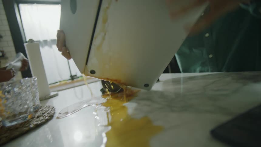 A man typing on a laptop attempts to lift his coffee mug but accidently spills it on his computer