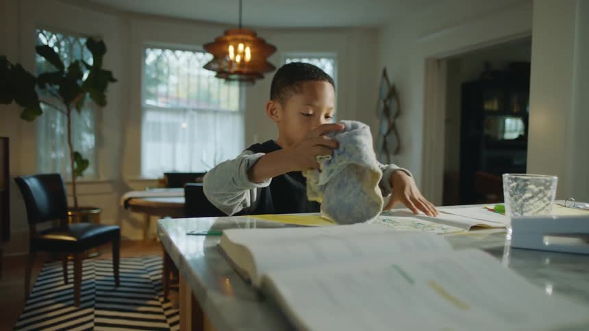 Boy drawing at counter spill juice then cleans it making a face with his mom when she joins him