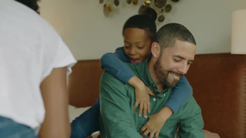 Boy smiles and back hugs father while father sits rubbing his foot after he stepped on toys