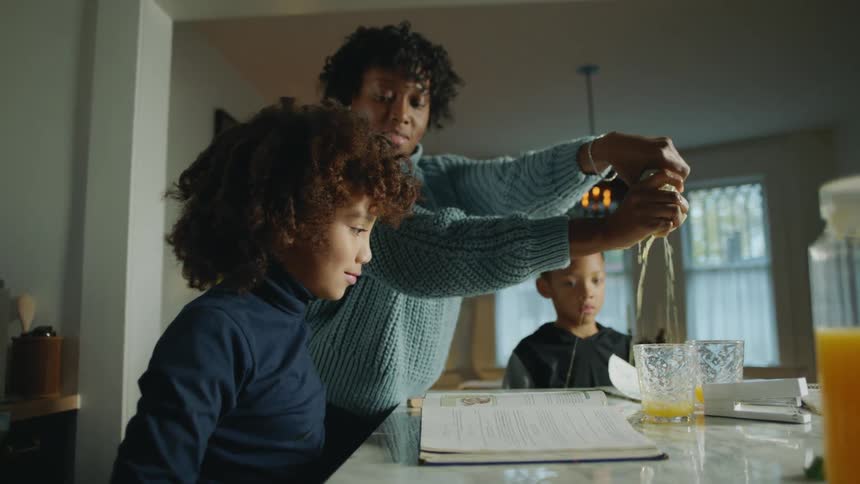 A woman wipes up a spilled drink from a table as two children are sitting at the table with books open