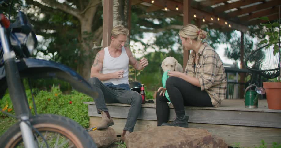 Blonde man with white undershirt gray pants and brown boots holds beer bottle and pets brown dog with blonde woman wearing plaid shirt and black leggings sitting on country house porch