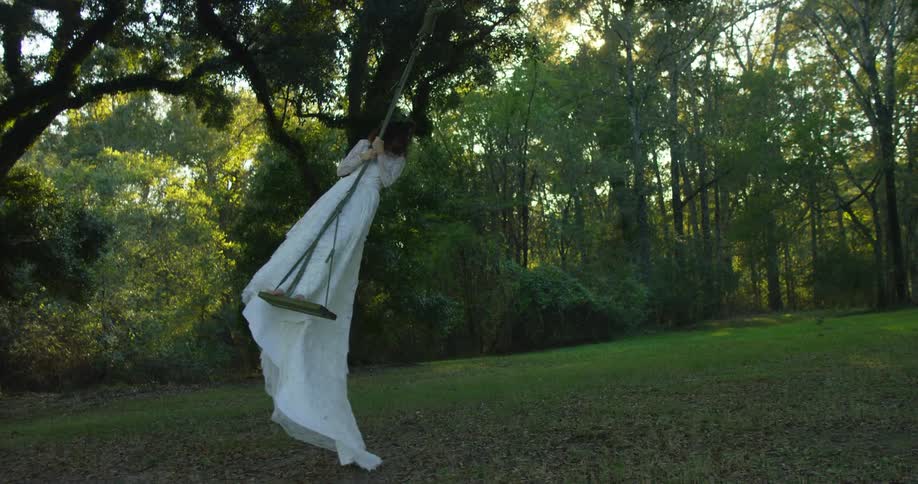 Woman wearing wedding dress stands on swing and swings across field covered in fallen leaves