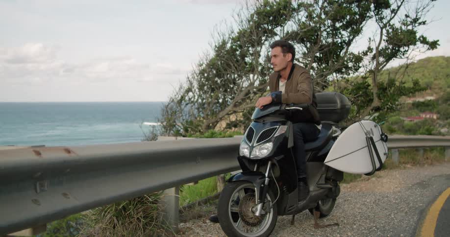 Young man sits over a parked motorcycle at the side of a road near the ocean