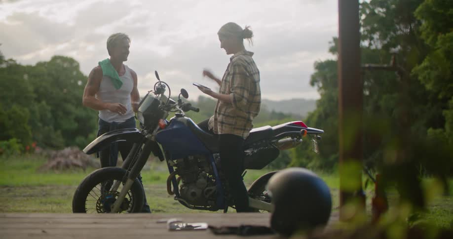 A man is repairing his motorcycle as a woman shows him her phone