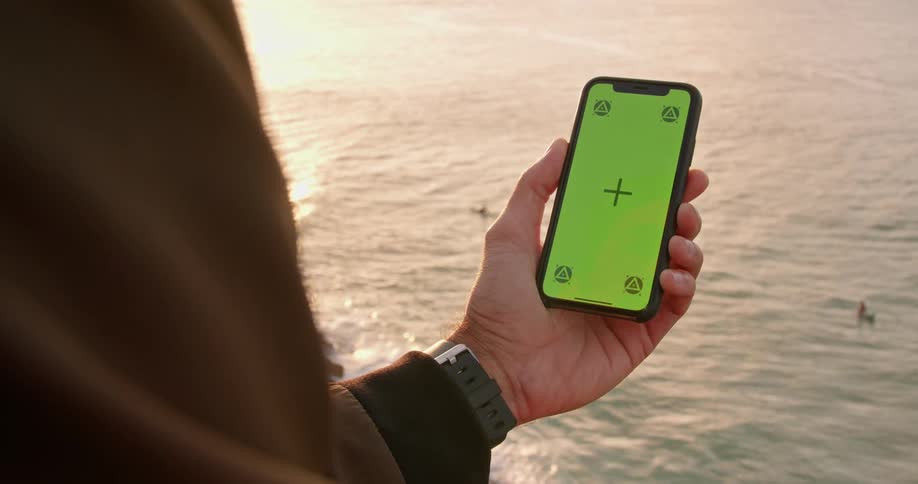 Man holds mobile phone looking down as surfers ride waves near water shining bright with reflected sunlight