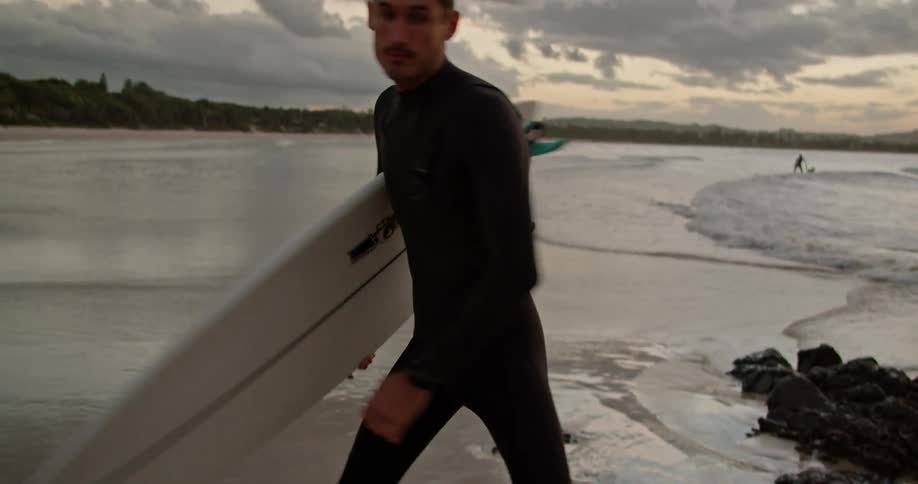 Male surfer with wetsuit holds surfboard warms up on the beach at dusk