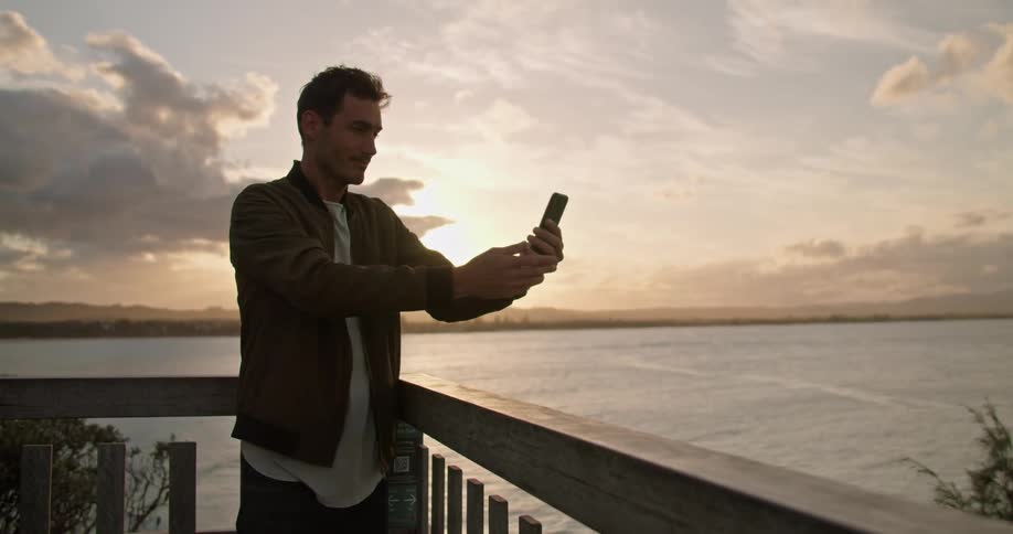 A man is standing on a deck with a large lake in the background while he video chats on his smartphone at dusk