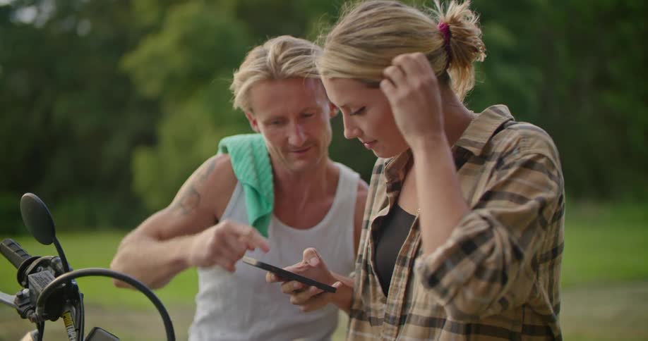 A man is sitting on a motorcycle talking to a woman who is showing him something on her cell phone