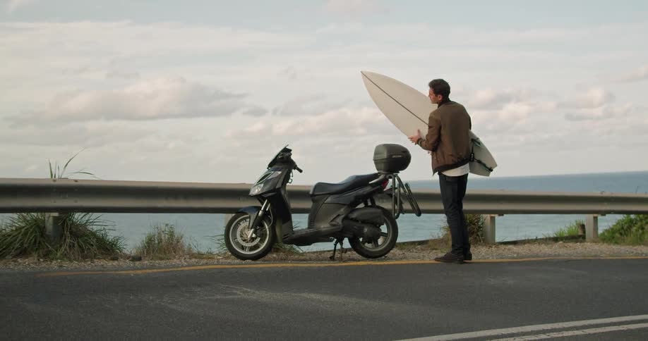 A man removes a surfboard from a carrier on the side of a motorcycle parked on the side of a road and carries the surfboard out of frame
