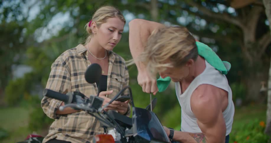 Woman sits on motorcycle talking to man as he works on the motorcycle