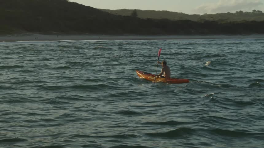 An aerial view of a man paddling a canoe across a body of water toward a beach at dusk