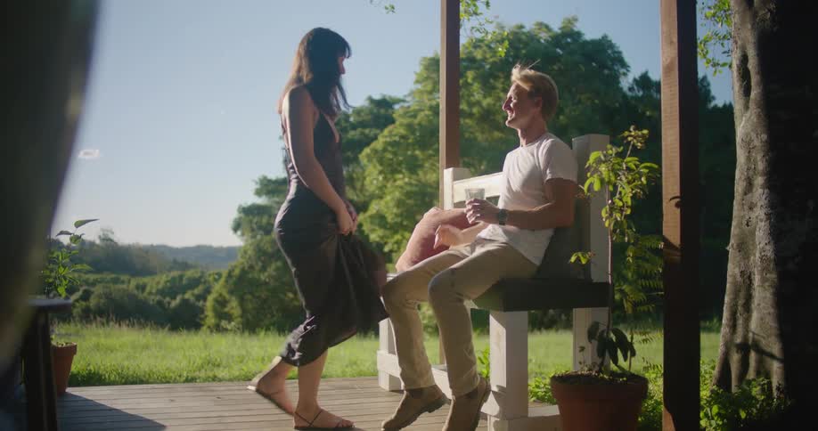 Blonde man with white t shirt and beige pants sits on bench with woman with brown wavy hair and dress in the garden kissing each other